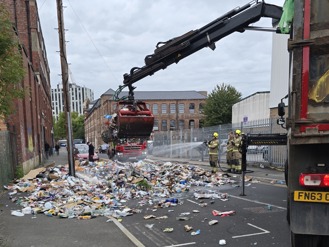 Firefighters spray water onto a large pile of mixed recycling that has been emptied onto a city street after a fire started inside the collected recycling. A grab arm from a waste truck moves the recycling material to find the cause of the fire. Another fire engine and several buildings line the background.